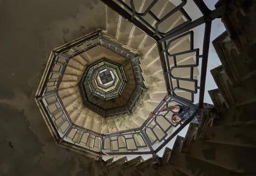 This architectural photograph showcases the spiral staircase inside the Volta Tower in Como, Italy. The image captures the staircase from a ground-up perspective, revealing its geometric and decorative railings that create a striking pattern as they ascend toward the top of the tower. Taken in the afternoon during the early autumn season, the photograph highlights the interplay of natural light streaming in from above, illuminating the stairwell’s textured surfaces. The Volta Tower itself serves as a recognizable landmark in Como, and the spiral staircase is a notable feature within this historic Italian structure.